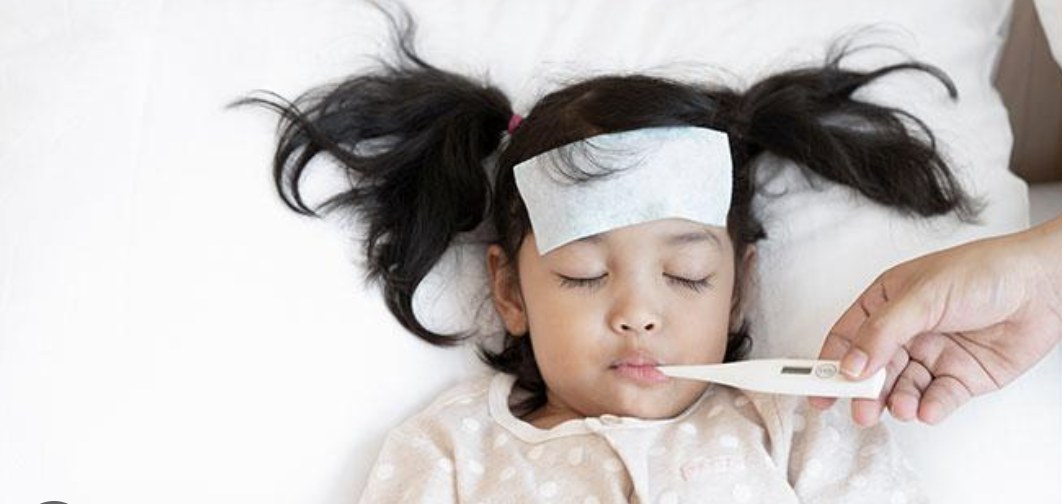 Child with fever resting in bed while a parent checks her temperature